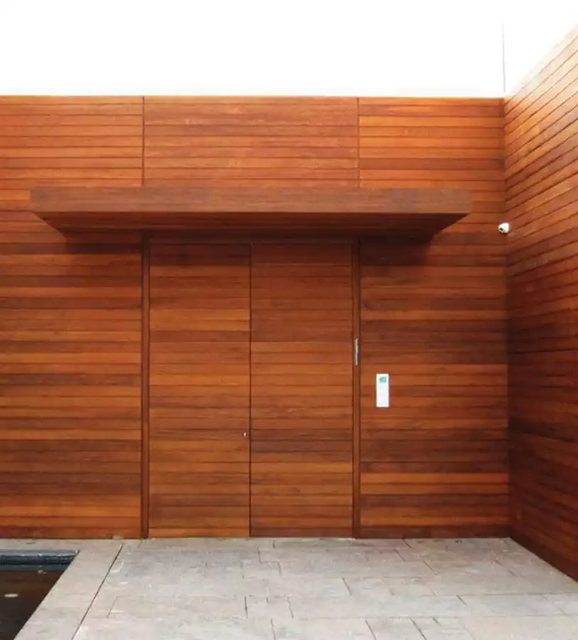 דלת כניסה מחופה בסרגלי עץ, נטמעת בחיפוי קיר החוץ. 'דלתות יסמין'.